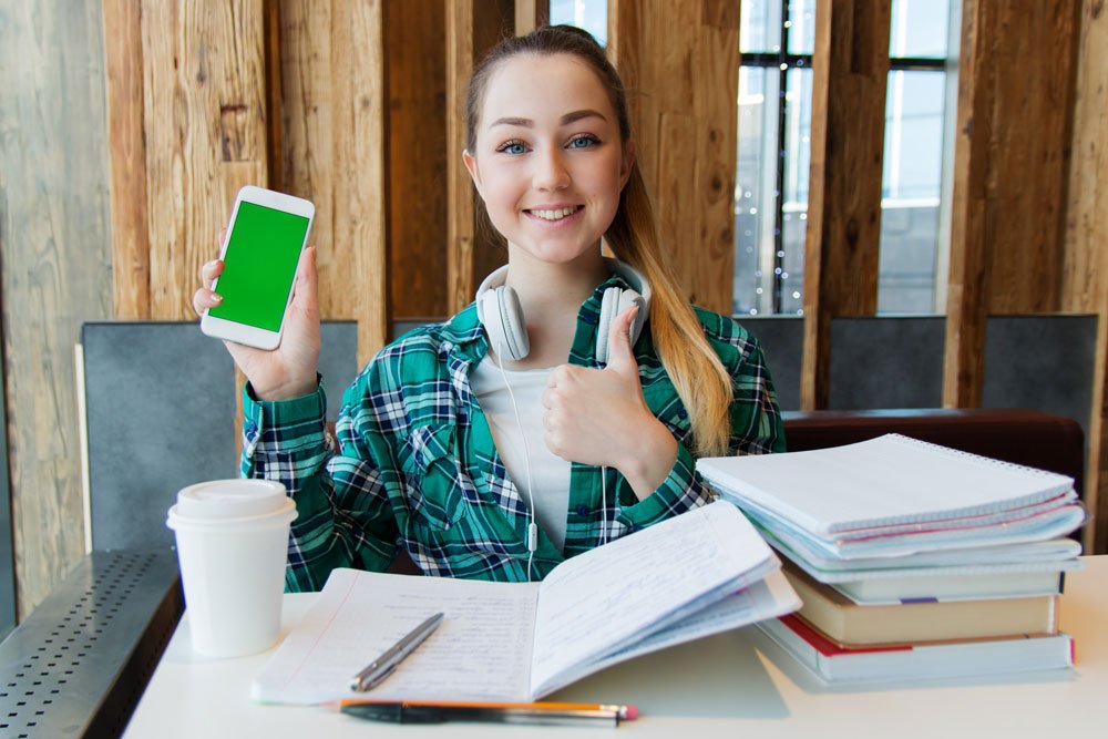 young student with large pile of books doing homework while listening to podcasts